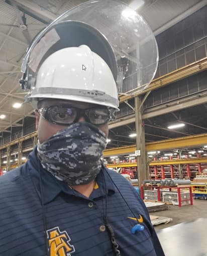 A man wearing a hardhat with face shield, safety glasses and a gaiter looks at the camera.